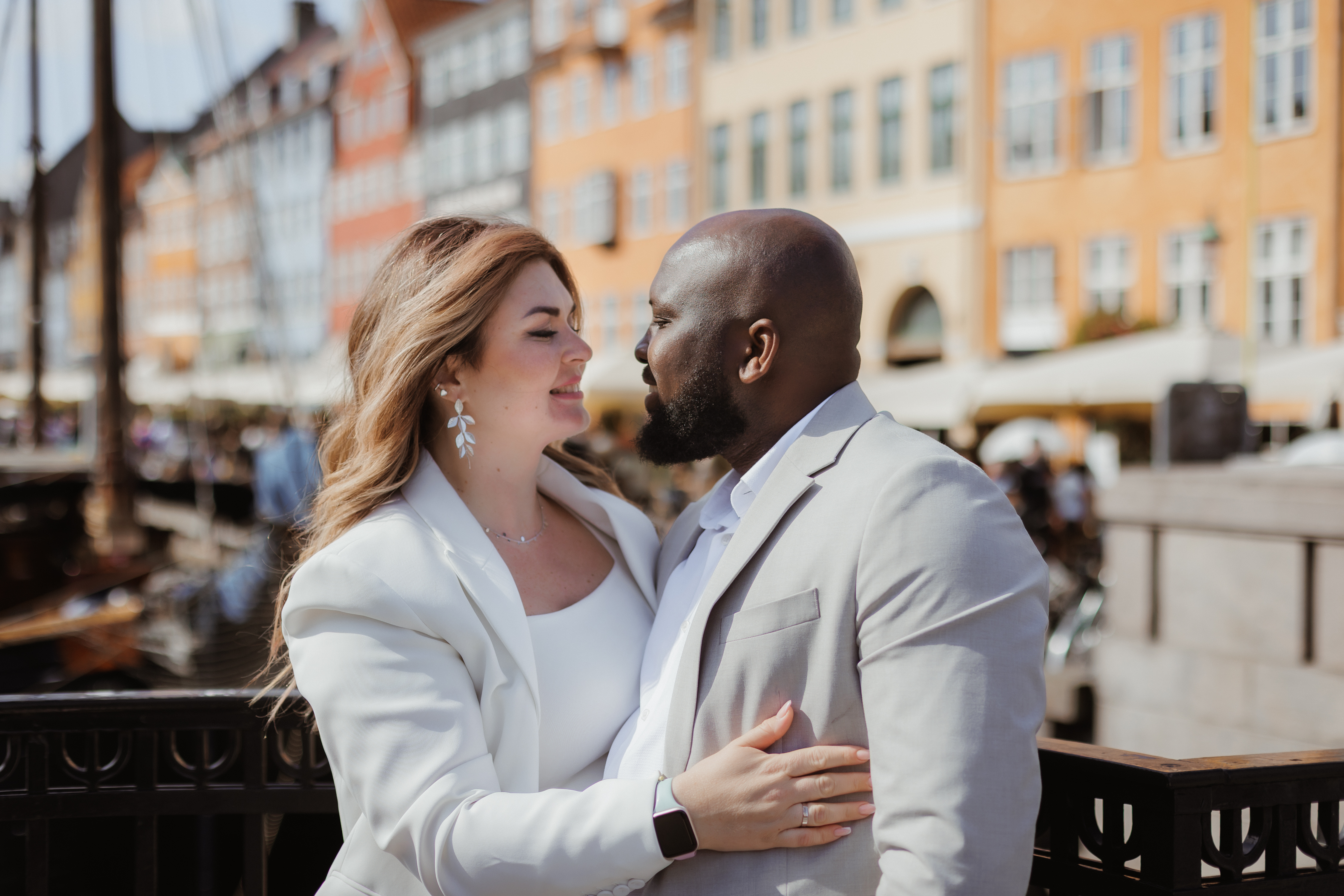 Ein dunkelhäutiger Herr und eine hellhäutige Dame, festlich für eine Trauung gekleidet, schauen sich vor einer Häuserzeile am Pier tief in Augen.